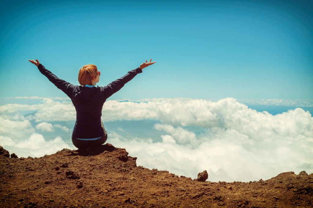 A woman enjoys a scenic view atop a cliff in Kula, Hawaii, surrounded by clouds and blue sky.